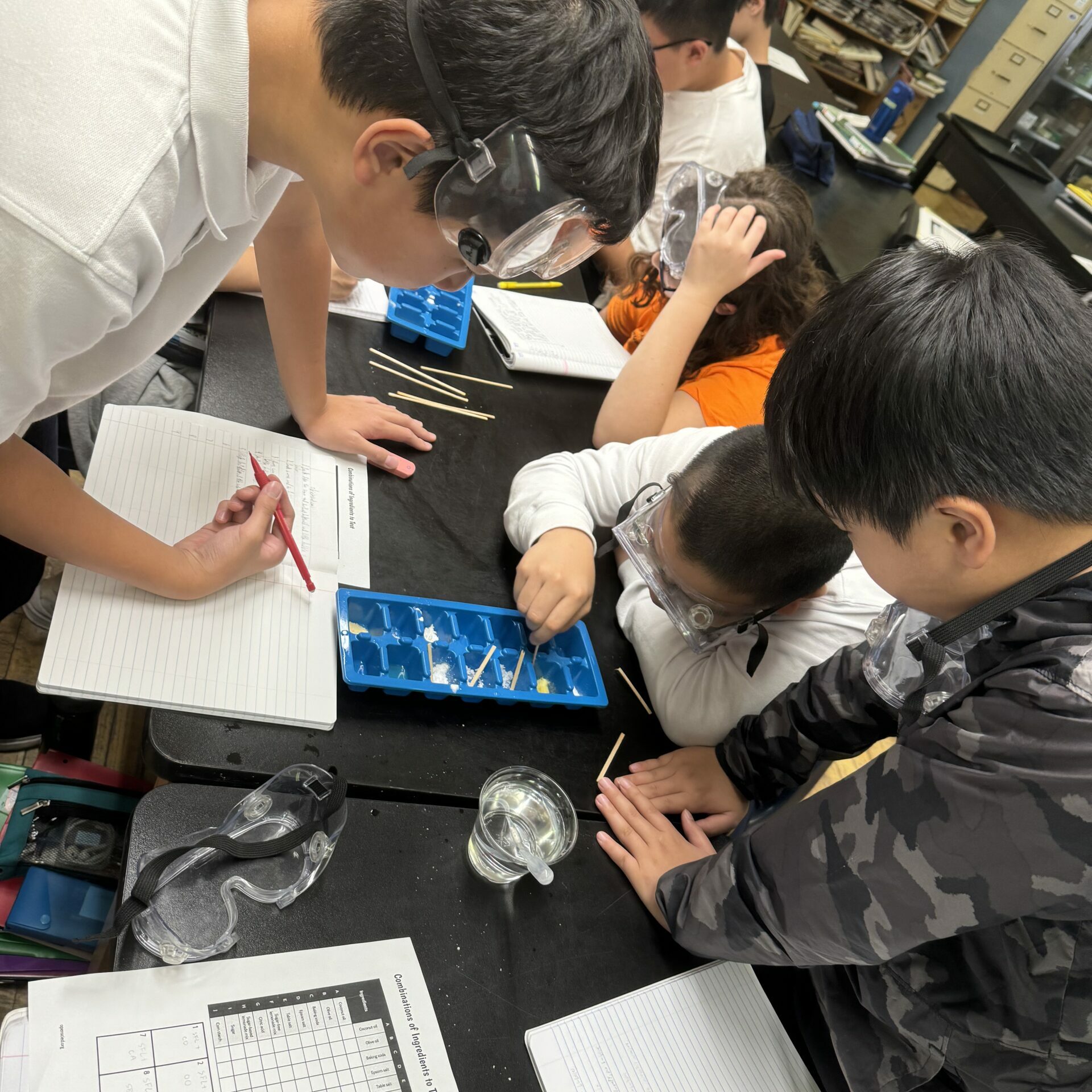 Students wearing safety goggles work together during a science lab activity, mixing materials in trays and recording observations in notebooks while a teacher supervises in the classroom.