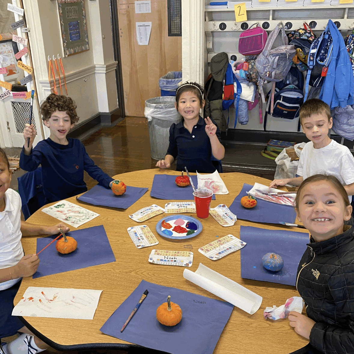 Five young students sit around a classroom table painting small pumpkins during an art activity. Paintbrushes, paint palettes, and paper are spread across the table as the students smile and show their work. Backpacks and coats hang on hooks along the classroom wall behind them.